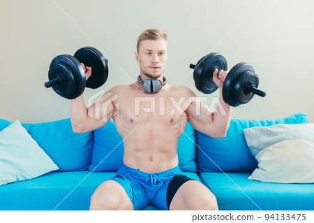 Young handsome athletic man trains with dumbbells, doing sports at home, sitting on sofa listening to music on headphones, looking at the camera Young handsome athletic man trains with dumbbells, doing sports at home, sitting on sofa listening to music on headphones, looking at the camera 94133475