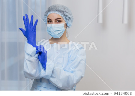 Young female doctor prepares for surgery, wears blue surgical gloves, in a white coat and mask, looks at the camera Young female doctor prepares for surgery, wears blue surgical gloves, in a white coat and mask, looks at the camera 94134068