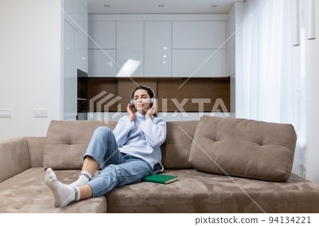 Young beautiful female student at home sitting on sofa listening to audiobooks in headphones, studying, learning a foreign language, preparing for exams Young beautiful female student at home sitting on sofa listening to audiobooks in headphones, studying, learning a foreign language, preparing for exams 94134221