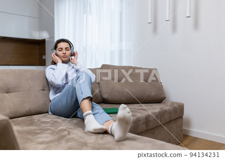 Young beautiful female student at home sitting on sofa listening to audiobooks in headphones, studying, learning a foreign language, preparing for exams Young beautiful female student at home sitting on sofa listening to audiobooks in headphones, studying, learning a foreign language, preparing for exams 94134231