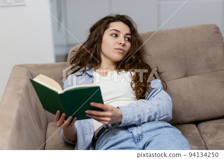 Young beautiful woman lying at home on the couch and reading a book, resting, looking at the camera, smiling 94134250