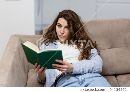 Portrait of a young beautiful female student lying on the couch at home, reading a book, studying, preparing for an exam, looking at the camera, smiling Portrait of a young beautiful female student lying on the couch at home, reading a book, studying, preparing for an exam, looking at the camera, smiling 94134251