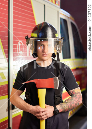Photo of fireman wearing helmet with ax against fire engine. Image of young man firefighter with helmet in head against background of fire truck 94137502