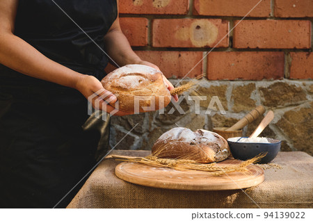 Midsection of a baker on black chef's apron holding a loaf of fresh baked whole grain sourdough wheaten bread 94139022