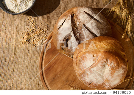 Top view of loaves of fresh homemade bread on a wooden board with scattered grains and wheat spikelets on the sackcloth 94139030