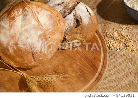 Variety of loaves of freshly baked bread on a wooden board with scattered grains and wheat ears on a burlap tablecloth 94139031