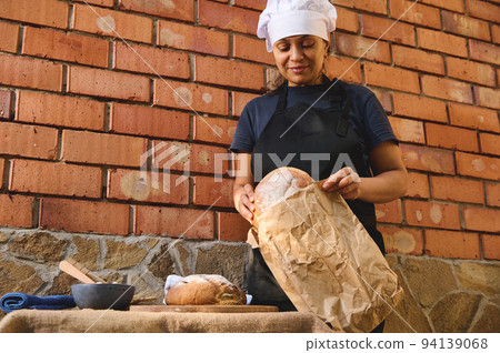 Charming woman baker, shop assistant in artisan bakery shop, packing fresh sourdough wheaten bread in eco bag, for sale 94139068