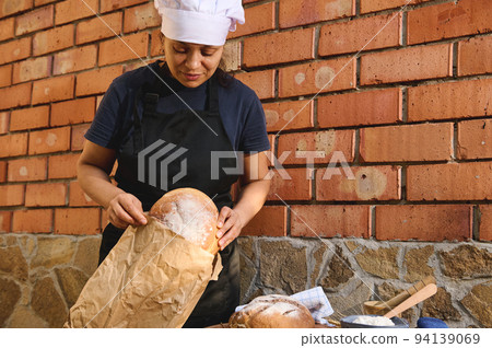 Beautiful woman baker, shop assistant in artisan bakery shop, packing fresh sourdough wheaten bread in eco bag, for sale 94139069