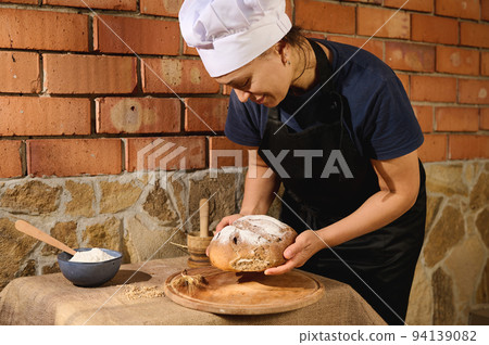 Inspired pretty woman baker in black chefs uniform stands over brick wall and holds a loaf of fresh baked wheaten bread Inspired pretty woman baker in black chefs uniform stands over brick wall and holds a loaf of fresh baked wheaten bread 94139082