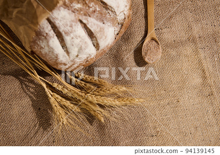 A loaf of whole grain bread sprinkled with flour, spikelets of wheat and a wooden spoon with bran on sacklock tablecloth 94139145