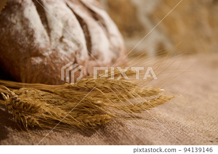 Selective focus on spikelets of wheat and blurred homemade whole grain freshly baked bread on table covered with burlap 94139146