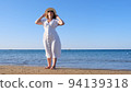 mature woman in sunglasses in a straw hat and white dress walks along the blue sea coast on a sunny 94139318