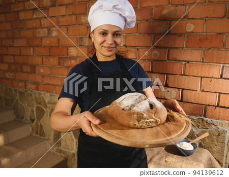 Cute woman baker smiles at camera, holding a wooden board with fresh baked whole grains sourdough bread with wheat ears Cute woman baker smiles at camera, holding a wooden board with fresh baked whole grains sourdough bread with wheat ears 94139612