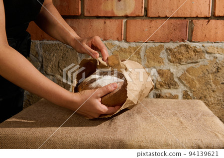 Selective focus on the hands of baker putting healthy fresh homemade whole grain bread into an eco recyclable paper bag Selective focus on the hands of baker putting healthy fresh homemade whole grain bread into an eco recyclable paper bag 94139621