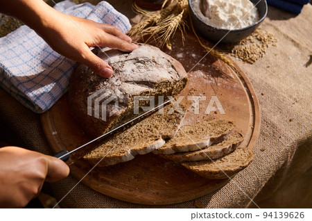 View from above of hands cutting a homemade whole grain round loaf of healthy sourdough rye bread on a wooden board. 94139626