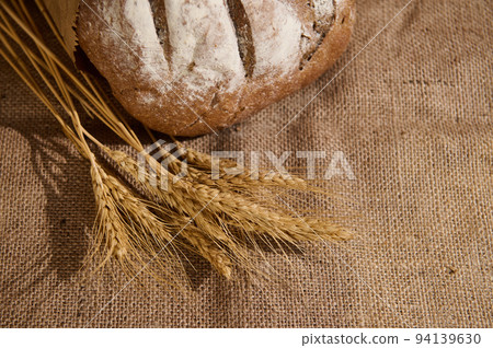 Selective focus on the ears of wheat next to a craft authentic homemade fresh baked whole grain bread on burlap surface 94139630