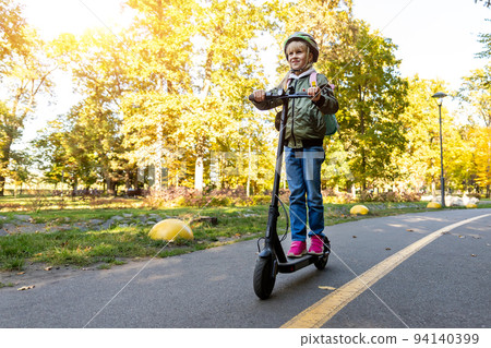 Profile view portrait of cute blond little caucasian school girl wear helmet enjoy having fun riding electric scooter city street park outdoors on sunny day. Healthy sport children activities outside Profile view portrait of cute blond little caucasian school girl wear helmet enjoy having fun riding electric scooter city street park outdoors on sunny day. Healthy sport children activities outside 94140399