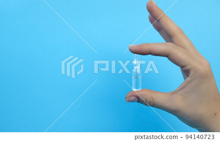 Close-up of a hand holding an ampoule with a vaccine or medicine on a blue background. Covid-19 prevention concept. Close-up of a hand holding an ampoule with a vaccine or medicine on a blue background. Covid-19 prevention concept. 94140723