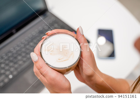 Close-up of beautiful female hands holding a large white cup of cappuccino. A business woman sits in a cafe working at a computer. Close-up of beautiful female hands holding a large white cup of cappuccino. A business woman sits in a cafe working at a computer. 94151161