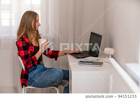European professional woman sitting with laptop at home office desk, positive woman studying while working on PC. She is wearing a red plaid shirt and jeans. European professional woman sitting with laptop at home office desk, positive woman studying while working on PC. She is wearing a red plaid shirt and jeans. 94151233