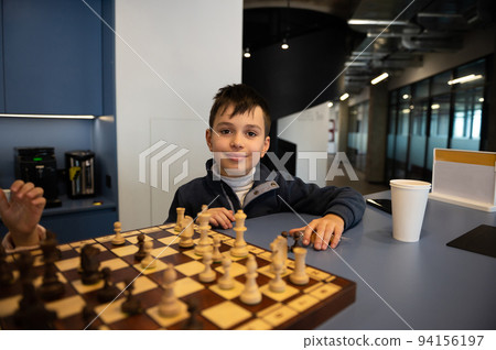 Charming smart dark-haired Caucasian child sitting at a chessboard and cute smiles looking at camera while playing chess Charming smart dark-haired Caucasian child sitting at a chessboard and cute smiles looking at camera while playing chess 94156197