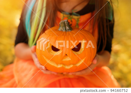 A child holds a pumpkin with a carved sinister smile in his hands at the sunset of the autumn sun. Traditions on Halloween. close-up A child holds a pumpkin with a carved sinister smile in his hands at the sunset of the autumn sun. Traditions on Halloween. close-up 94160752