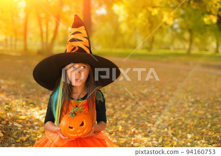 A surprised girl dressed as a witch in a black hat with an orange ribbon holds a sinister pumpkin in her hands on Halloween day. trick or treat. sales season. 94160753