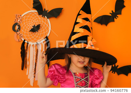 A girl on an orange studio background puts on a witch hat for Halloween, hiding her face under it. Cute shy kid. Halloween concept. A girl on an orange studio background puts on a witch hat for Halloween, hiding her face under it. Cute shy kid. Halloween concept. 94160776