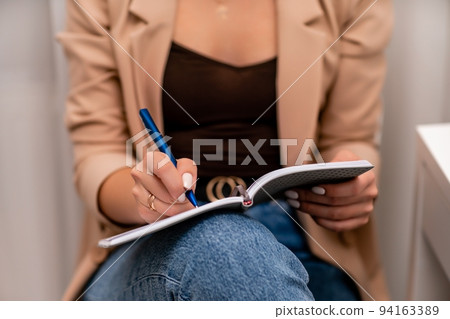 European woman writes in a notebook. She is wearing a beige jacket and jeans. European woman writes in a notebook. She is wearing a beige jacket and jeans. 94163389