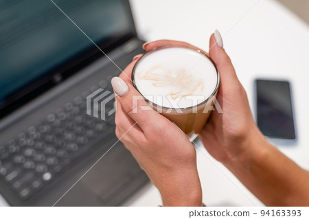 Close-up of beautiful female hands holding a large white cup of cappuccino. A business woman sits in a cafe working at a computer. Close-up of beautiful female hands holding a large white cup of cappuccino. A business woman sits in a cafe working at a computer. 94163393