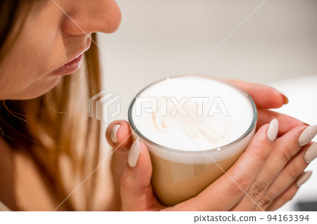 Close-up of beautiful female hands holding a large white cup of cappuccino. A woman is sitting in a cafe. 94163394