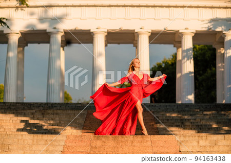 A woman in a long red dress against the backdrop of sunrise, bright golden light of the sun's rays. The concept of femininity, harmony. 94163438