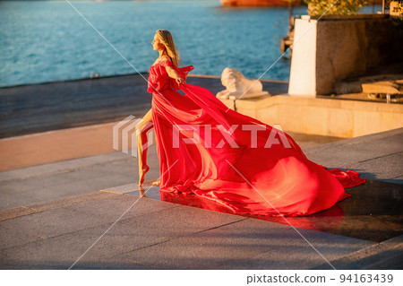 A woman in a long red dress against the backdrop of sunrise, bright golden light of the sun's rays. The concept of femininity, harmony. 94163439