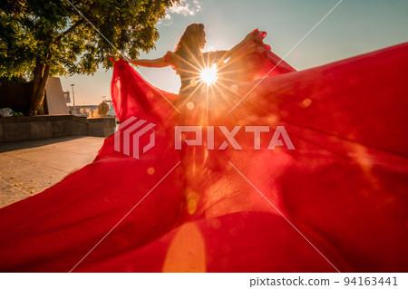 A woman in a long red dress against the backdrop of sunrise, bright golden light of the sun's rays. The concept of femininity, harmony. 94163441
