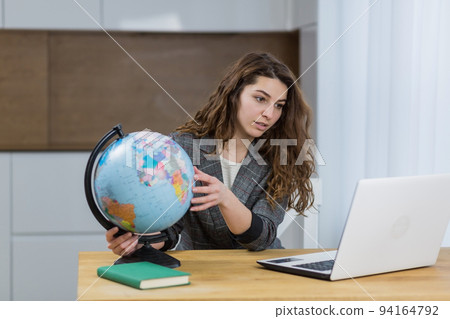 Young female teacher, travel agency worker teaches online from laptop, shows globe, tells via video call Young female teacher, travel agency worker teaches online from laptop, shows globe, tells via video call 94164792