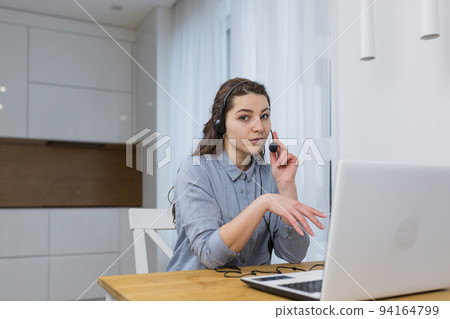 Young beautiful woman, call center employee communicates online with laptop and headphones with customers, provides advice, at home at the table, quarantine 94164799