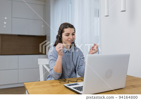 Young beautiful woman, call center employee communicates online with laptop and headphones with customers, provides advice, at home at the table, quarantine 94164800