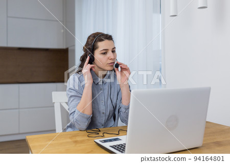 Young beautiful woman, call center employee communicates online with laptop and headphones with customers, provides advice, at home at the table, quarantine 94164801