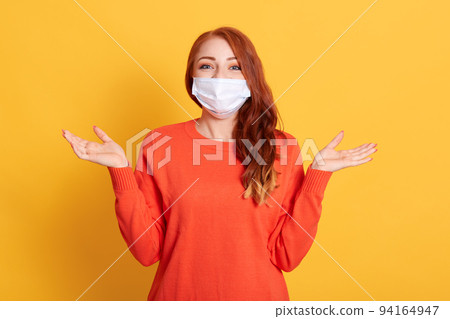 Young red haired woman in protective medical mask spreading hands aside, female wearing orange casual sweater posing isolated over yellow background. Young red haired woman in protective medical mask spreading hands aside, female wearing orange casual sweater posing isolated over yellow background. 94164947