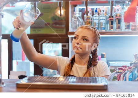 Woman bartender with glass of beer at work, red haired barman with pigtails wearing white casual t shirt tapping drink, looking at tap, posing with showcase with alcohol on background. Woman bartender with glass of beer at work, red haired barman with pigtails wearing white casual t shirt tapping drink, looking at tap, posing with showcase with alcohol on background. 94165034