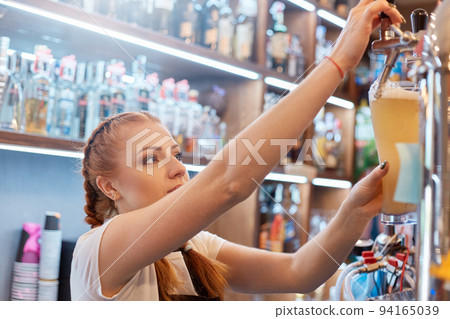 Pretty lady bartender with glass of beer at work, barkeeper tapping alcohol drink, red haired waiter at counter with showcase with alcohol on background. 94165039
