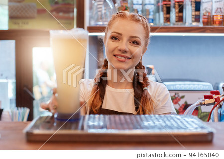 Red haired positive woman with pigtails holding beer glass while working at bar, attractive bartender wearing white t shirt and apron, stands with showcase with booze. 94165040