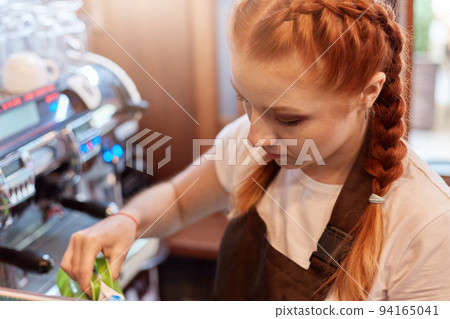Red haired barista working in cafe, looks concentrated, posing in white t shirt and brown apron with coffee machine on background, girl with pigtails in coffee shop. 94165041