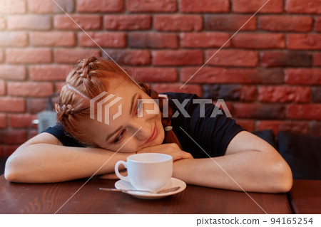 Beautiful woman with cup of coffee lies on table and looking smiling aside, tired student has break, posing against brick wall in black wall, red haired lady resting in cafeteria. Beautiful woman with cup of coffee lies on table and looking smiling aside, tired student has break, posing against brick wall in black wall, red haired lady resting in cafeteria. 94165254