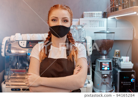 Woman in small cafe wearing medical mask at counter in cafe, keeps hands folded, looks at camera, wearing white t shirt, brown apron, black protective mask, protects from virus. Woman in small cafe wearing medical mask at counter in cafe, keeps hands folded, looks at camera, wearing white t shirt, brown apron, black protective mask, protects from virus. 94165284