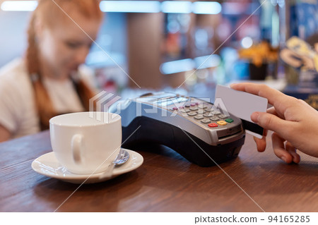 Customer paying for cup of coffee with card, using terminal for paying, white cup of coffee and red haired barista on background, faceless person uses device and card. Customer paying for cup of coffee with card, using terminal for paying, white cup of coffee and red haired barista on background, faceless person uses device and card. 94165285