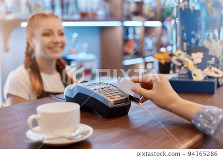 Customer paying through mobile phone over electronic reader in coffee shop, faceless customer holding card, smiling red haired waitress over counter. Customer paying through mobile phone over electronic reader in coffee shop, faceless customer holding card, smiling red haired waitress over counter. 94165286