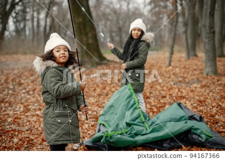 Two little black girls in tent camping in the forest. Two little sisters are puting up a tent in autumn forest. Black girls wearing khaki coats and beige hats. Two little black girls in tent camping in the forest. Two little sisters are puting up a tent in autumn forest. Black girls wearing khaki coats and beige hats. 94167366