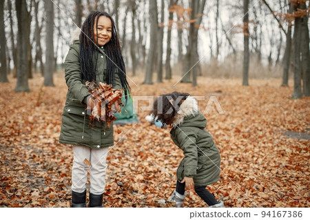 Beautiful little black girls standing near tent in the forest. Two little sisters playing with yellow leaves in autumn forest. Black girls wearing khaki coats. 94167386