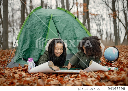 Beautiful little black girls in tent camping in the forest. Two little sisters lying in a tent in autumn forest and looking on a map. Black girls wearing khaki coats. Beautiful little black girls in tent camping in the forest. Two little sisters lying in a tent in autumn forest and looking on a map. Black girls wearing khaki coats. 94167396